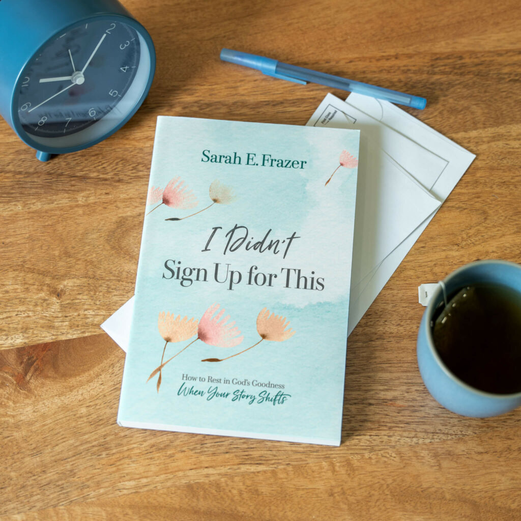 The book 'I Didn't Sign Up for This' by Sarah Frazer, resting on a table with a blue clock and coffee mug nearby, symbolizing reflection on life's challenges and discovering faith amidst adversity.