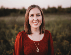 woman with light brown hair and a wide smile wearing a red shirt looks at the camera. She is standing in a field on a cloudy day.